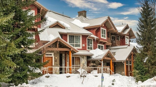 A large, multi-story wooden cabin with a snow-covered roof, surrounded by evergreen trees. - 2BR | Symphony | Dog Friendly | Hot Tub | Private Balconies | Village Location | Parking (Whistler)