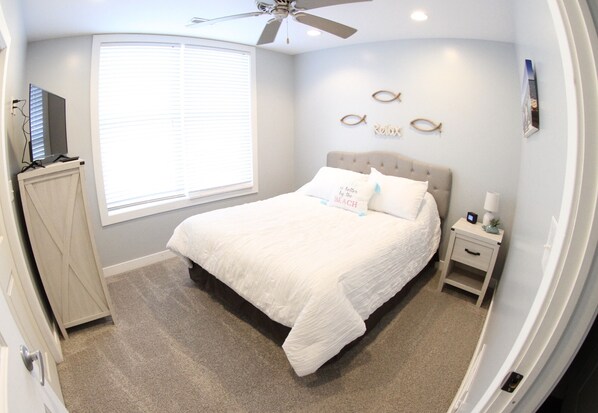 A bedroom with a bed, a nightstand, a ceiling fan, a window with blinds, and a wall-mounted TV.