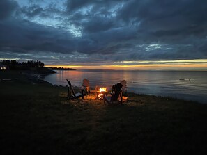 A beach scene with chairs and a fire, overlooking a calm sea and a sunset.