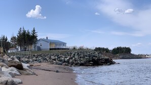 A beach with a rocky shoreline, a house with a deck, and trees in the background. - Oceanfront beach cottage panoramic views spectacular sunsets sandbars warm water (Pictou)