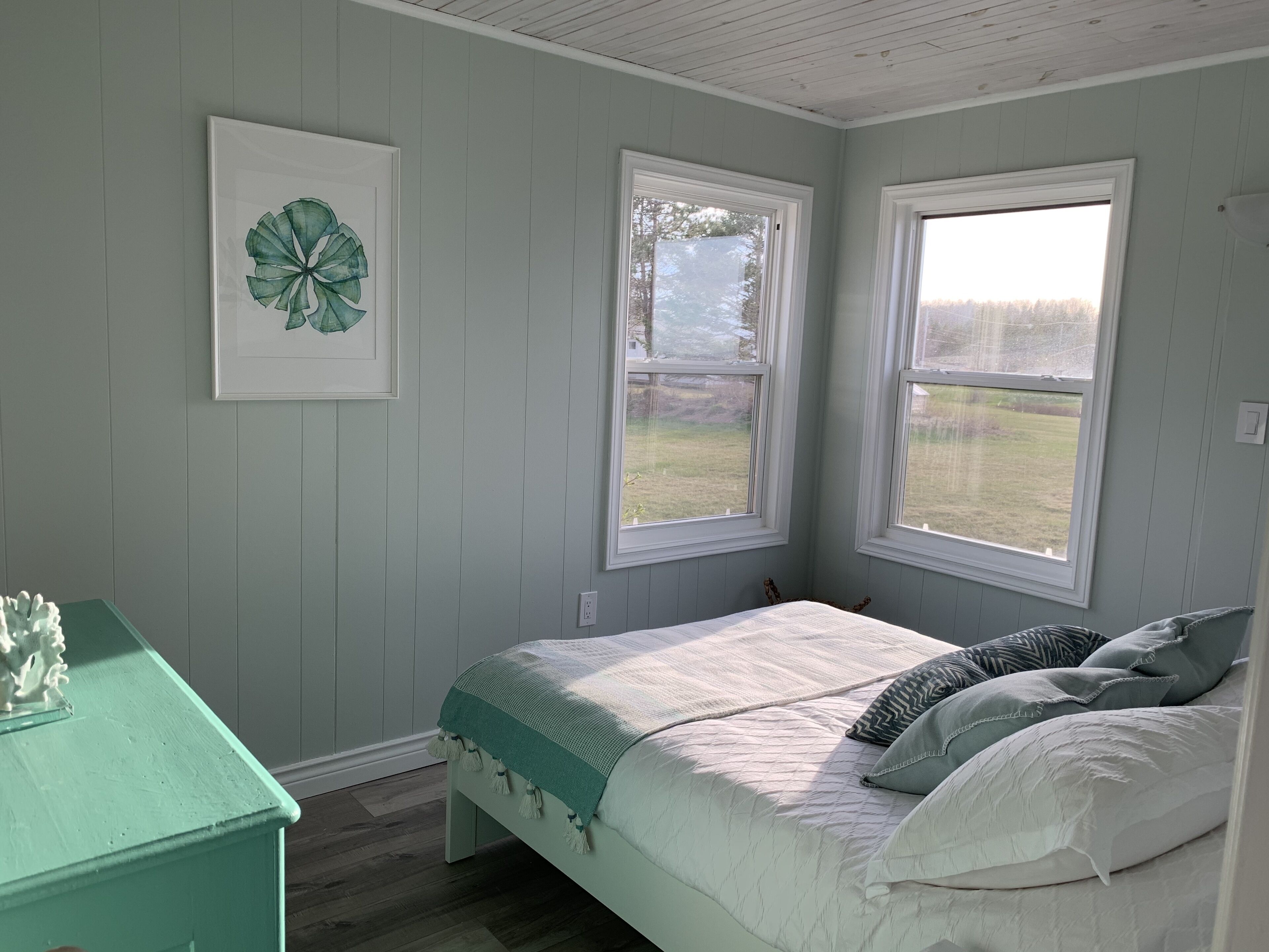 A bedroom with a bed, two windows, a wall-mounted picture, and a green dresser.