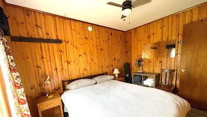 Une chambre à coucher aux murs lambrissés, comprenant un lit, une table de chevet, une lampe, un ventilateur et un miroir.