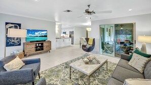 A spacious living room with a television, sofa, and armchair, a kitchen in the background, and a sliding glass door leading to an outdoor area.