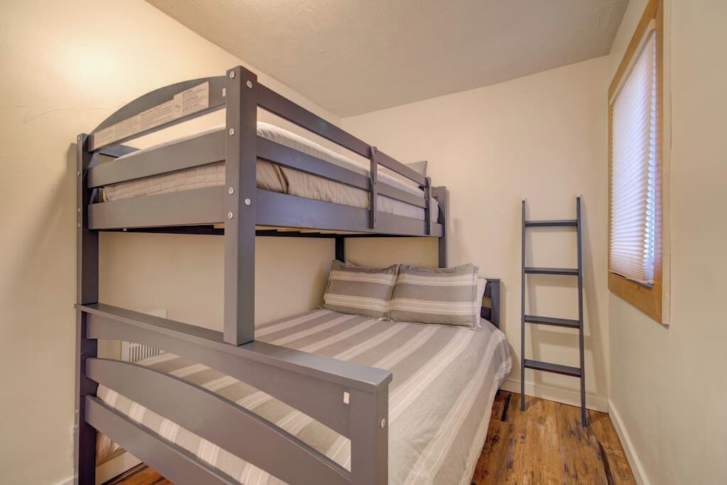 A bunk bed room with a ladder, wooden flooring, and a window with blinds.