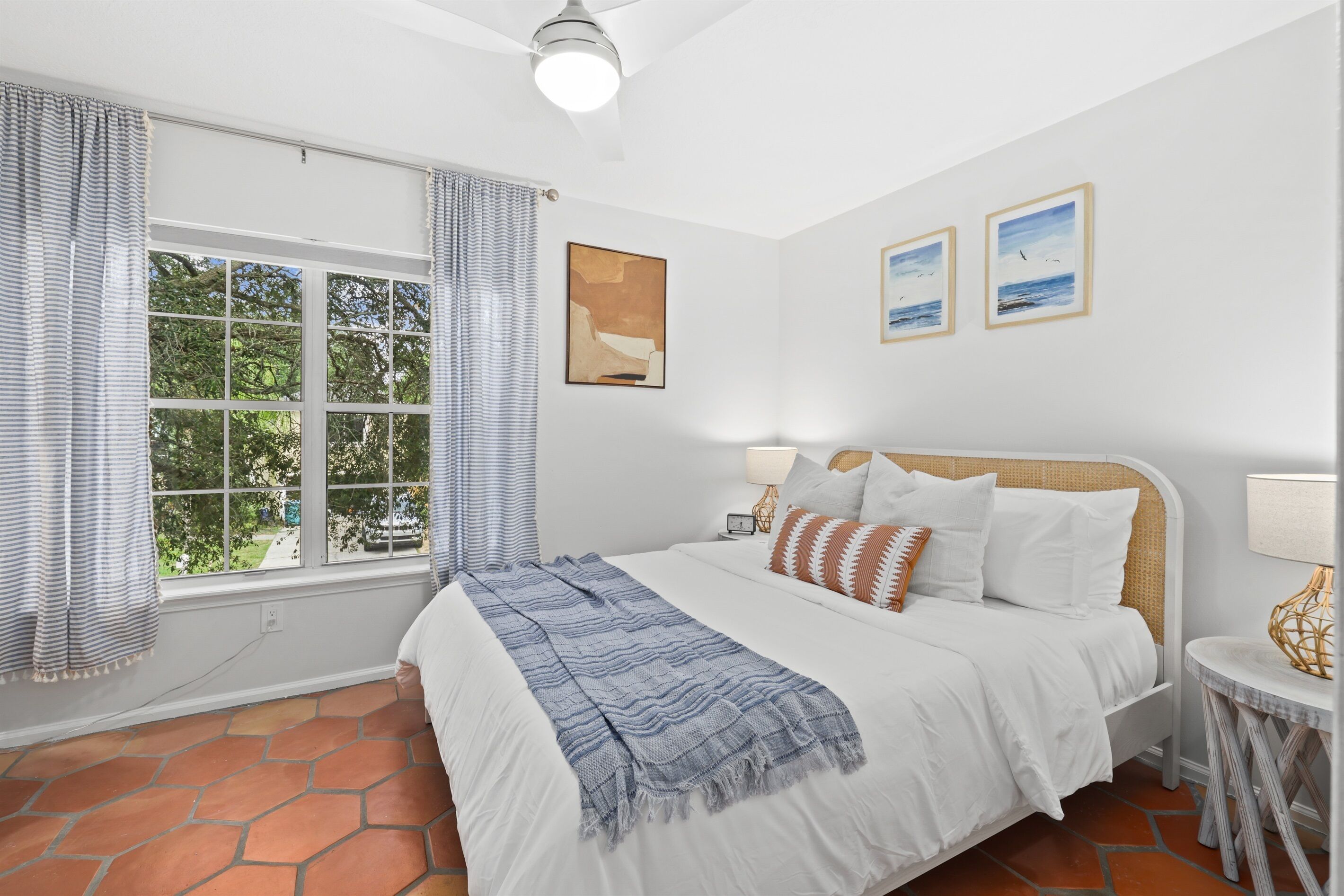 A bedroom with a large bed, a window with curtains, a ceiling fan, and artwork on the wall.