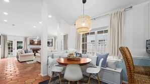 A modern living room with a round wooden table, white chairs, and a hanging light fixture. - Charming Retreat | Pool, Walk to Park & Coffee (Jacksonville Beach)