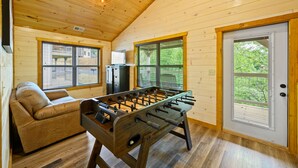 Habitación acogedora con techo de madera, un sofá, una mesa de fútbol de mesa y una puerta con vistas a la vegetación.