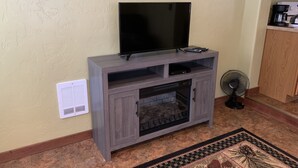 Uma sala de estar com televisão de tela plana sobre um suporte de madeira, lareira abaixo e um pequeno ventilador à direita.