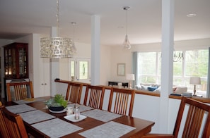 Ein Essbereich mit einem Holztisch, gedeckt für vier Personen, umgeben von Stühlen. Über dem Tisch hängt eine Chandelier, und zur rechten Seite ein Fenster mit Vorhängen.