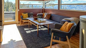 Een gezellig interieur van een chalet met een houten vloer, een bank, twee fauteuils, een eettafel en grote ramen met uitzicht op de bergen.