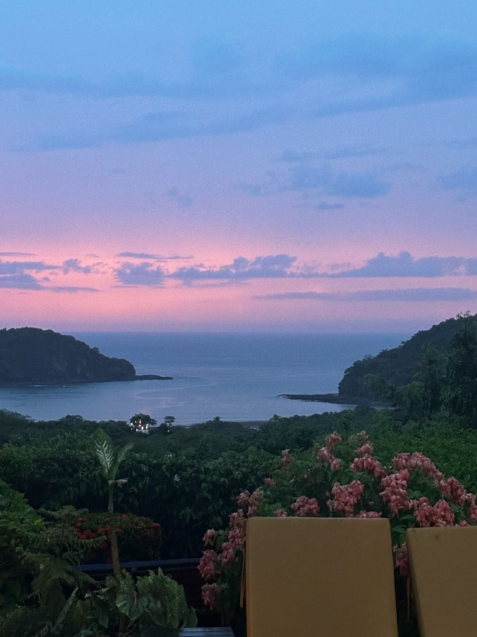 A coastal view with a sunset, greenery, and two empty chairs.