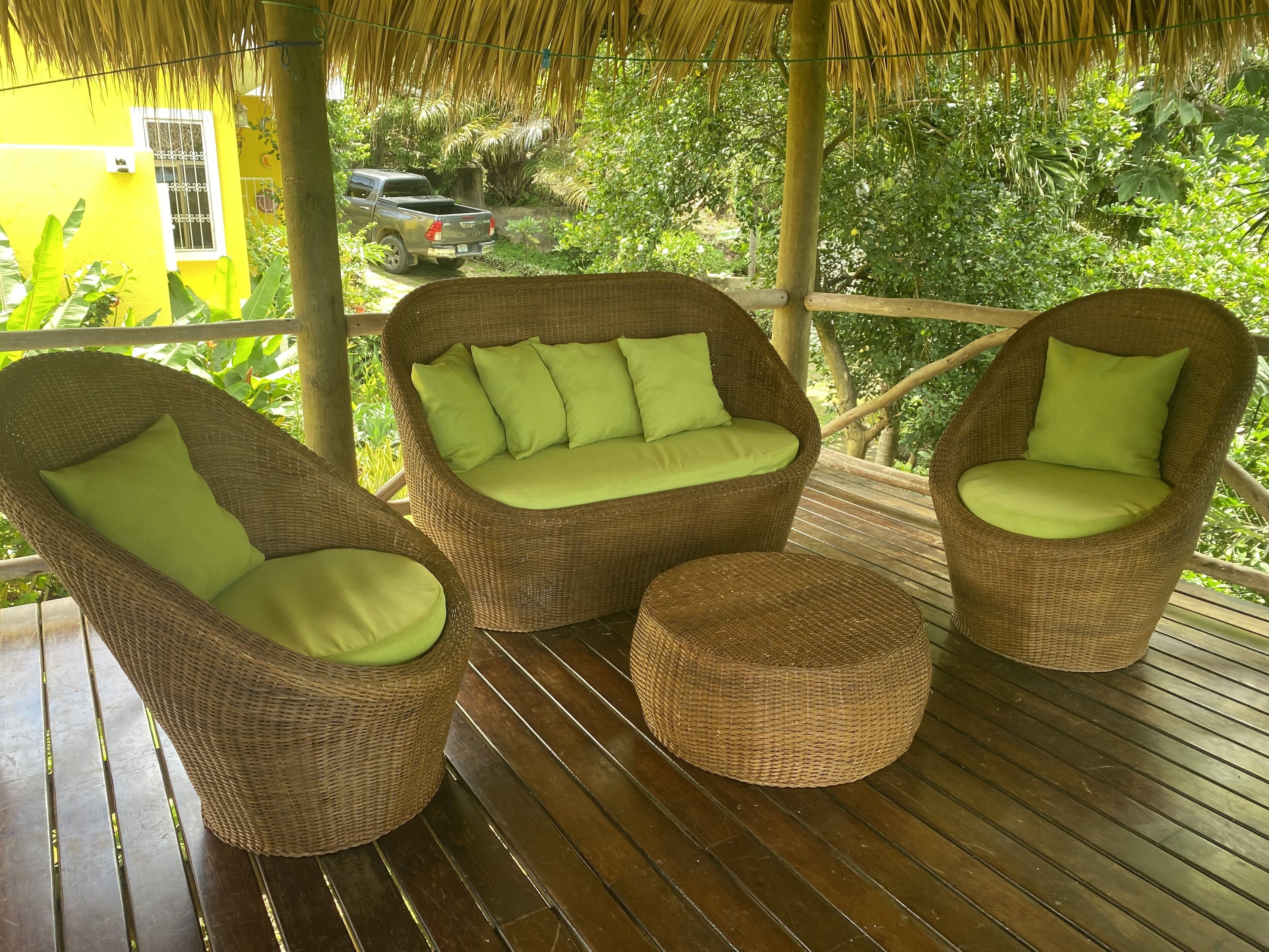 A covered outdoor seating area with wicker furniture and cushions, a wooden deck, and a view of a yellow building and parked vehicle in the background.