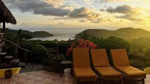 Eine Terrasse mit Liegestühlen, einem strohgedeckten Gebäude und Blick auf Meer und Hügel.