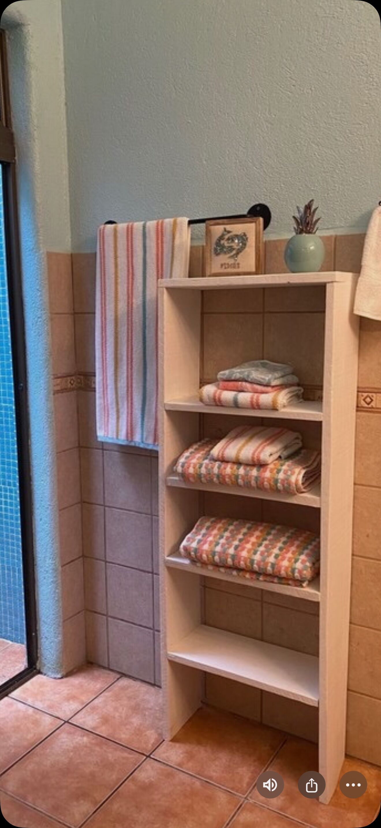 A bathroom with a wooden shelf holding folded towels and a striped towel hanging on the wall.