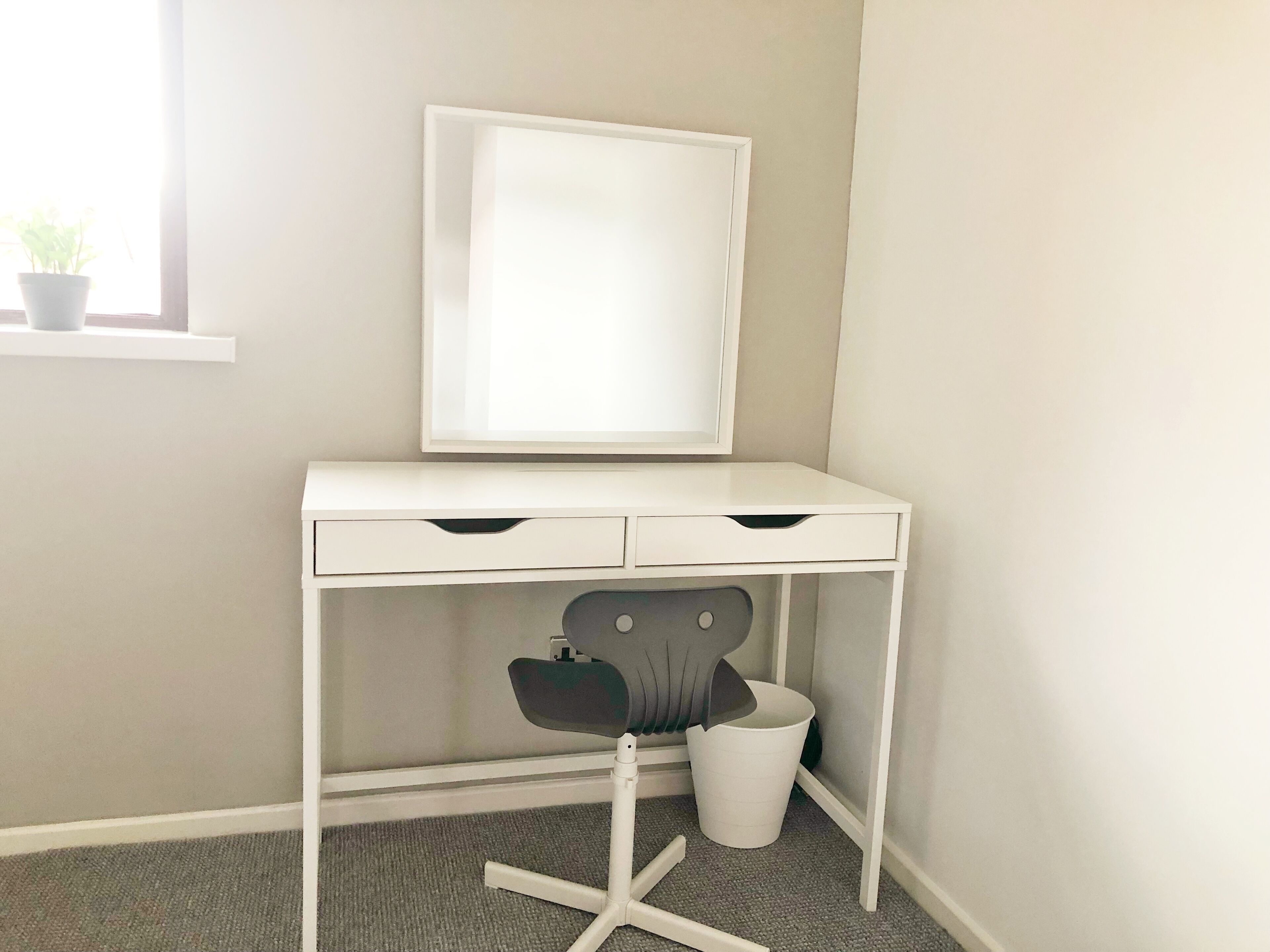 Een wit bureau met een spiegel, een stoel en een prullenbak in een hoek slaapkamer.