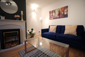 A living room with a blue sofa, a glass coffee table, a fireplace, and a painting on the wall.