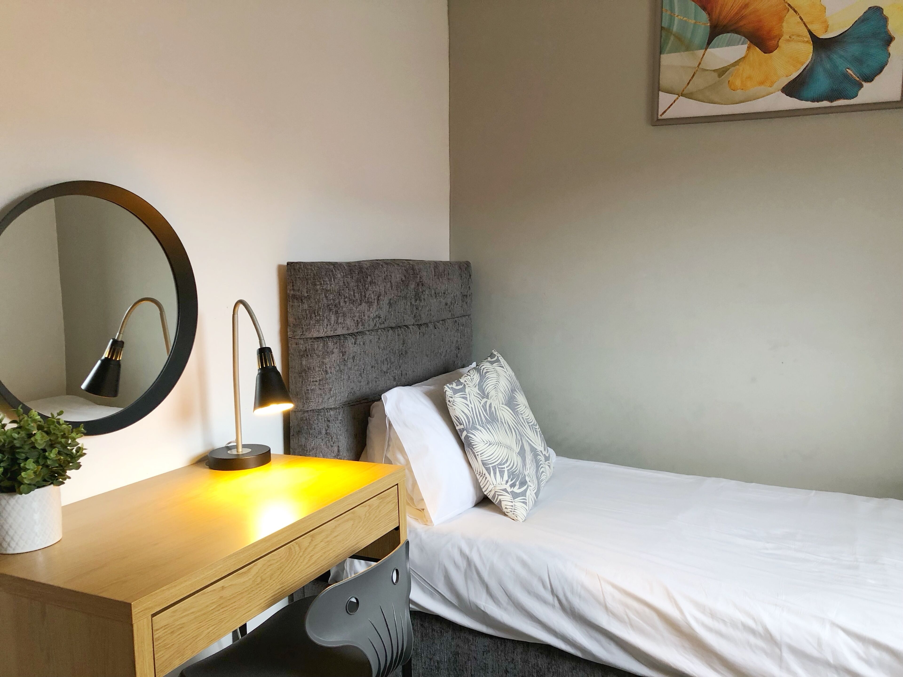 A bedroom with a bed, a bedside table with a lamp, a mirror, and a plant.