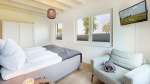 A bedroom with a bed, a grey sofa, a TV, and a window with blinds. - Pet friendly home in Büsum with sauna (Büsum)