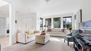 A modern living room with a sofa, ottoman, and armchair, a flat-screen TV, and large windows offering a view of the outdoors. - Family vacation home at Cultus Lake - Pet Friendly (Cultus Lake)