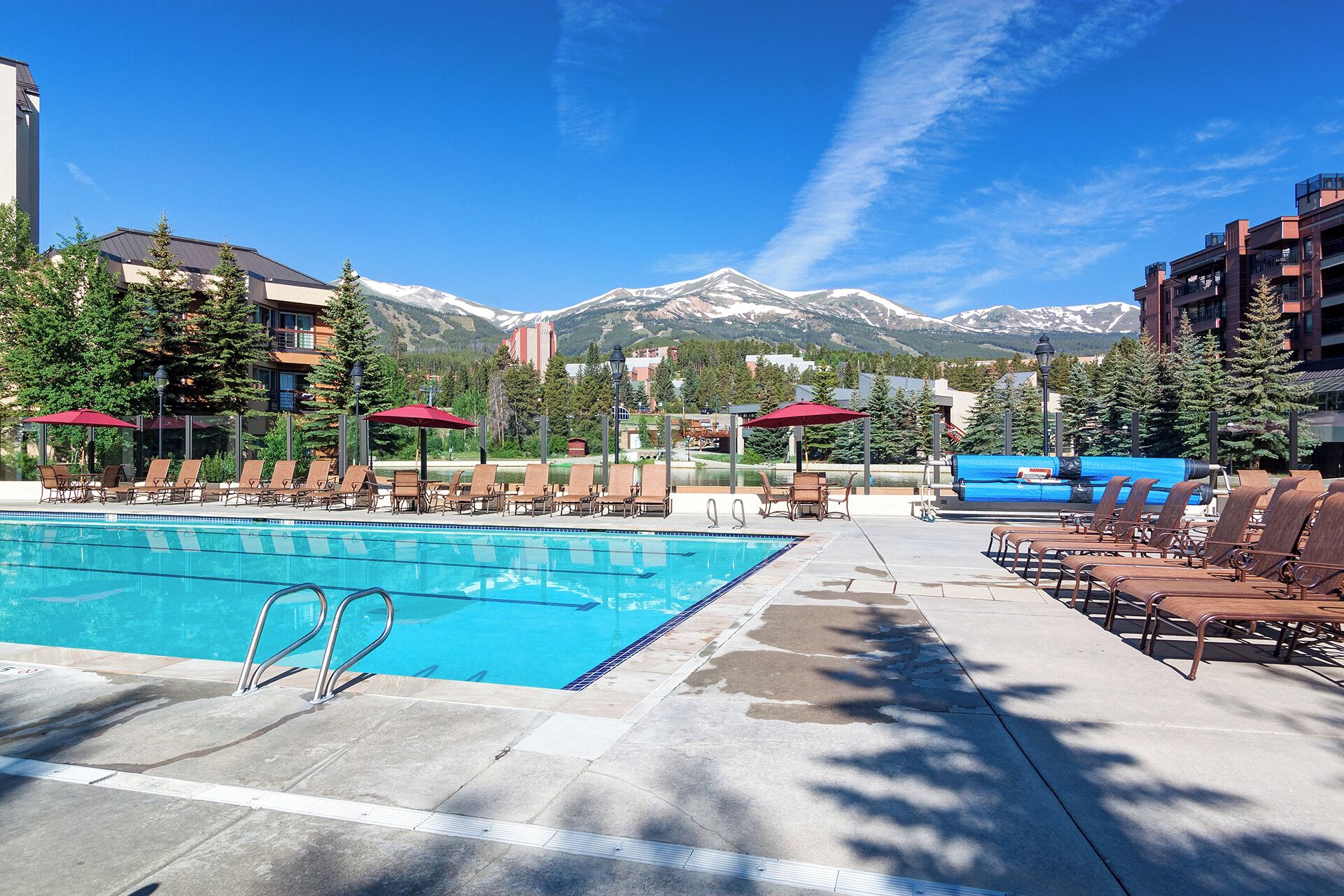Zona piscina con lettini e ombrelloni, circondata da edifici e montagne.