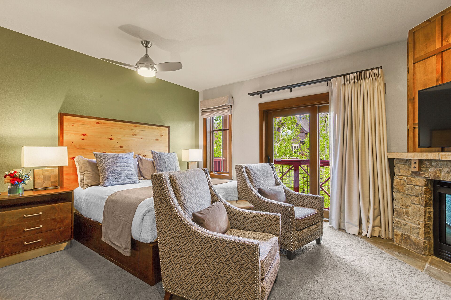 Une chambre à coucher comprenant un lit, deux fauteuils, une table de chevet, une lampe, un ventilateur de plafond, une fenêtre avec des rideaux et une cheminée.