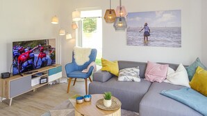 Ett vardagsrum med en tv-apparat som visar ett cykellopp, en blå fåtölj, en grå soffa med färgglada kuddar och en målning av en person vid havet.