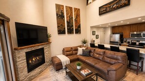 Una sala de estar con chimenea, un sofá modular, una mesa de centro y una cocina al fondo.