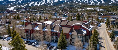 A multi-story building with a snow-covered roof, surrounded by a parking lot and trees, with a mountain range in the background.