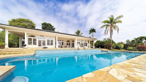 A large swimming pool with a covered patio area, surrounded by stone tiles and palm trees. - Hale Hilo Bay Tropical Hideaway (Hilo)