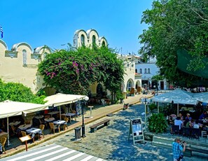 Un área de café al aire libre con mesas y sillas, un paso de peatones y un edificio con ventanas arqueadas al fondo.