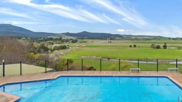A rectangular swimming pool with a wooden deck and black metal railing, surrounded by a grassy area and a fence, with a view of distant hills and a clear sky.