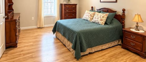 A bedroom with a wooden floor, a bed with a green bedspread, a dresser, a nightstand with a lamp, a window with curtains, and a wall-mounted picture.