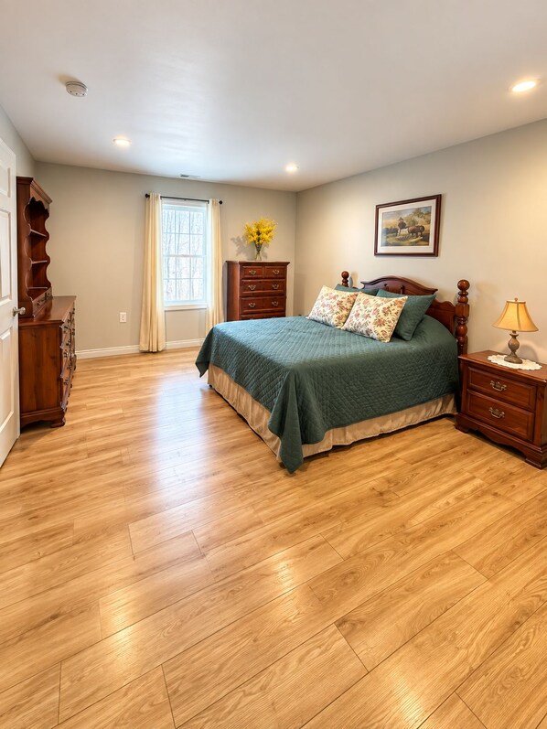 A bedroom with a wooden floor, a bed with a green bedspread, a dresser, a nightstand with a lamp, a window with curtains, and a wall-mounted picture.