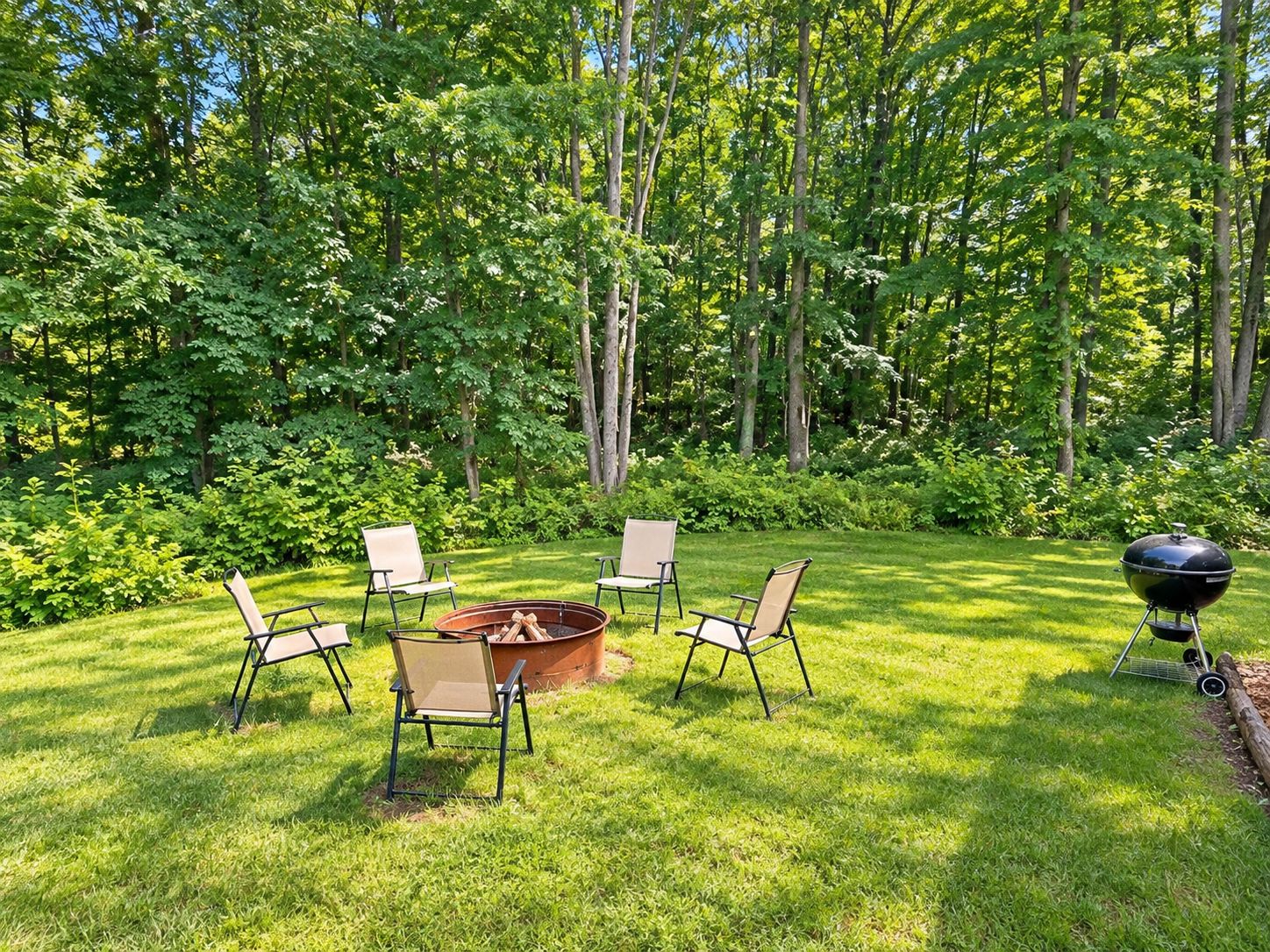A grassy outdoor area with a fire pit, four chairs, and a grill.
