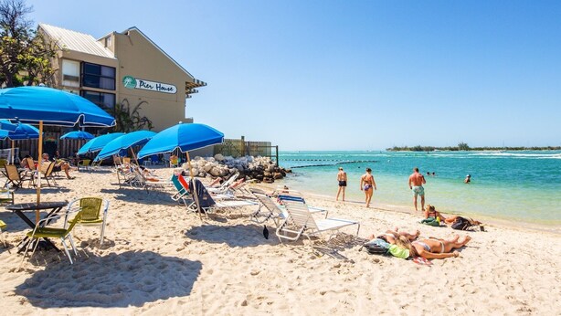 Ein Strand mit Liegestühlen, Sonnenschirmen und einem Gebäude im Hintergrund, auf dem 'Pier House' steht.