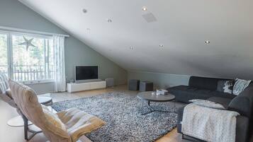 Uma sala de estar moderna com teto inclinado, uma janela grande, televisão de tela plana, sofá cinza, mesa de centro redonda e tapete estampado.