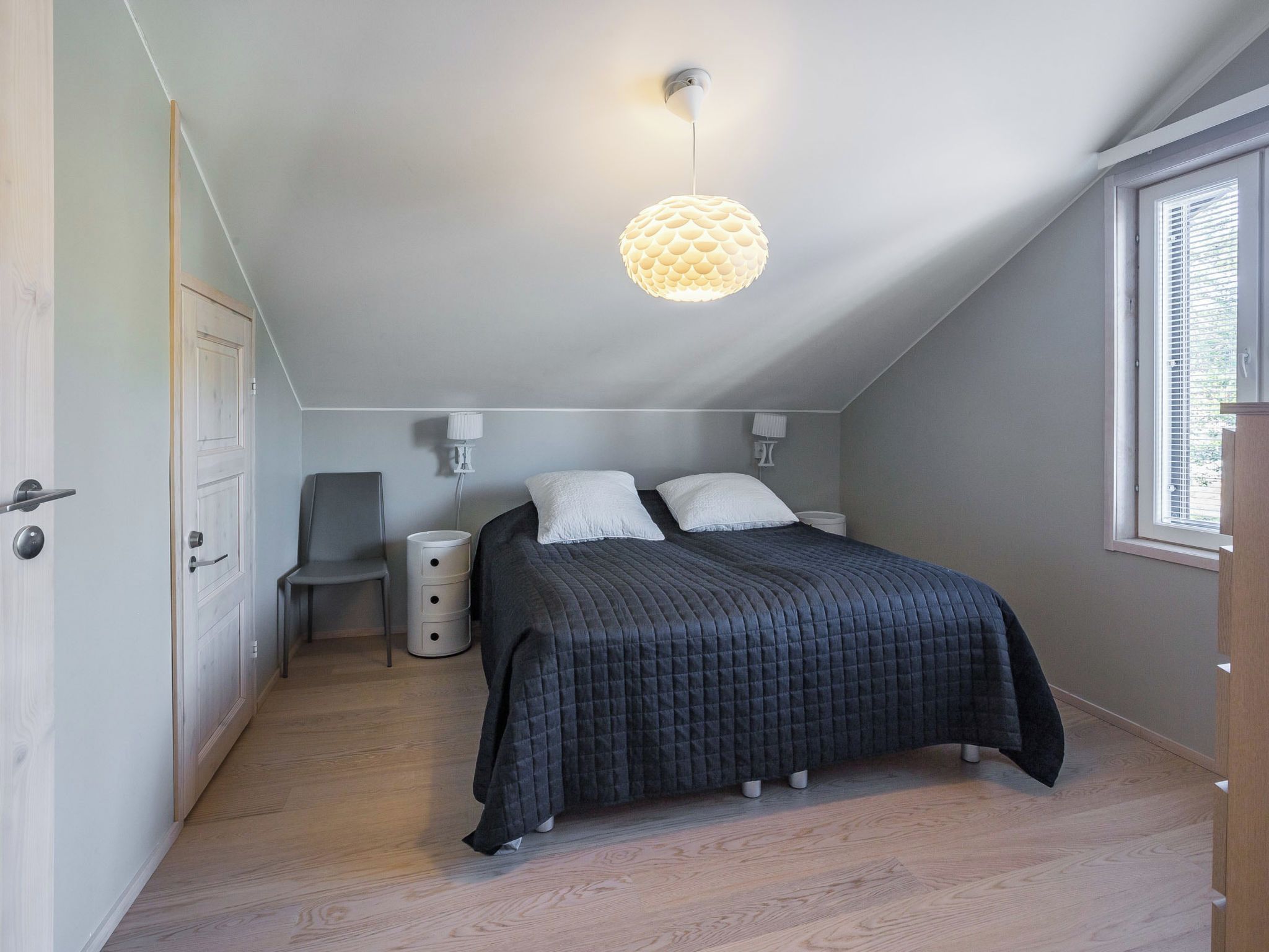 Une chambre avec un plafond en pente, un lit recouvert d’une couverture sombre, une chaise, une table de chevet et une fenêtre avec des stores.