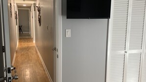 Un pasillo con piso de madera, un televisor de pantalla plana montado en la pared y una puerta blanca con persianas.