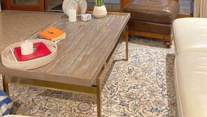 Ein Zimmer mit Holztisch, gepolstertem Sitzbrett, einem gemusterten Teppich und Blick auf Palmen durch eine Schiebetür.