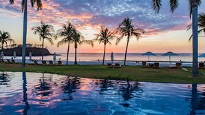 Blick auf ein Schwimmbecken mit Palmen, Liegestühlen und einem Sonnenuntergang über dem Meer.