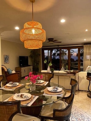A dining area with a glass table, wicker pendant light, and a view of palm trees through large windows.