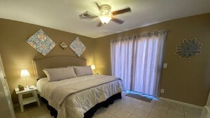 Un dormitorio con cama, mesita de noche, lámpara, ventilador de techo y cortinas.