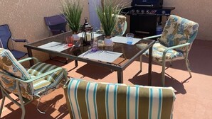 Un patio exterior con una mesa de vidrio, sillas a rayas, una parrilla y plantas en macetas.