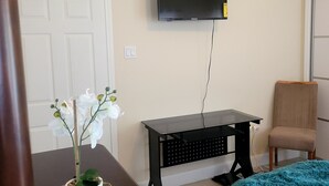 Una habitación con televisión montada en la pared, un escritorio con silla, una planta en maceta y una cama con manta azul.