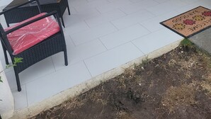 Un espacio de estar al aire libre techado con dos sillas de mimbre negro y un banco, una puerta blanca y una ventana con persianas.