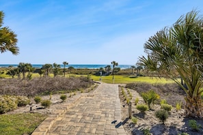 A paved pathway leading to a beach with palm trees and a clear view of the ocean.