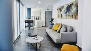 Una sala moderna con un sofá gris, una mesa de centro redonda y un reposapiés amarillo. Se ve una cocina al fondo con gabinetes blancos y una encimera negra.