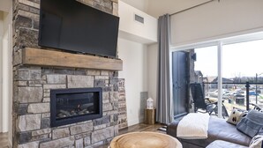 Ein modernes Wohnzimmer mit Kamin aus Stein, einem großen Flachbildfernseher, einer gemütlichen Sitzecke und Blick ins Freie.