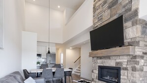 Ein modernes Wohnzimmer mit Kamin aus Stein, einem darüber hängenden Flachbildfernseher und einem Essbereich mit Tisch und Stühlen.