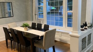 Un coin repas avec une table et des chaises en bois, une plante en pot et une vue sur la rue grâce à de grandes fenêtres.